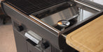 Close-up of a dark gray outdoor gas grill with a stainless steel interior, black control knobs, and a light wood side table attached.