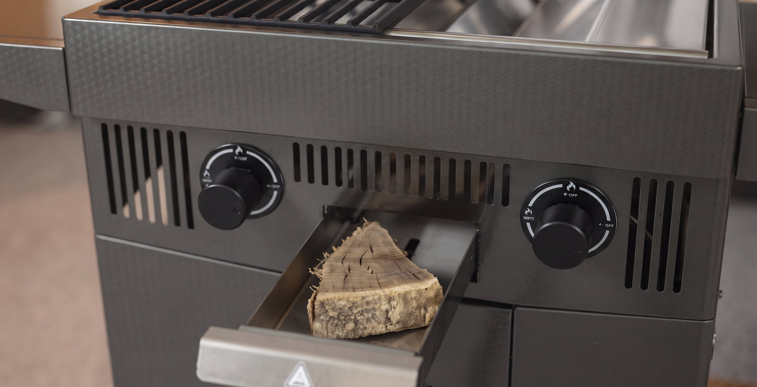 Close-up of a dark gray outdoor gas grill with two black control knobs and a wood slice inserted into the firebox opening.