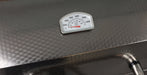 A close-up of a stainless steel grill lid featuring a white analog temperature gauge with black markings and a red liquid-filled expansion tube.