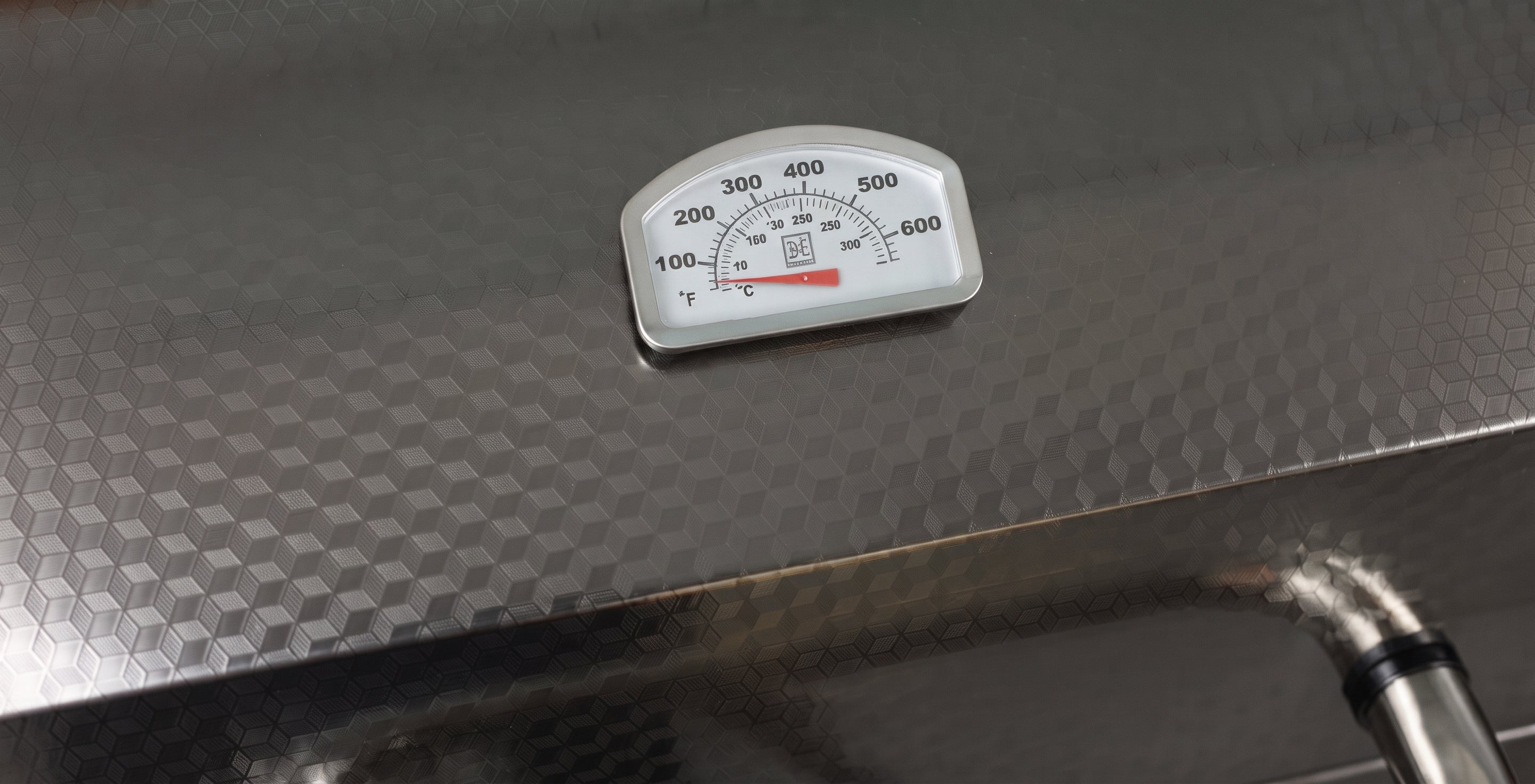 A close-up of a stainless steel grill lid featuring a white analog temperature gauge with black markings and a red liquid-filled expansion tube.