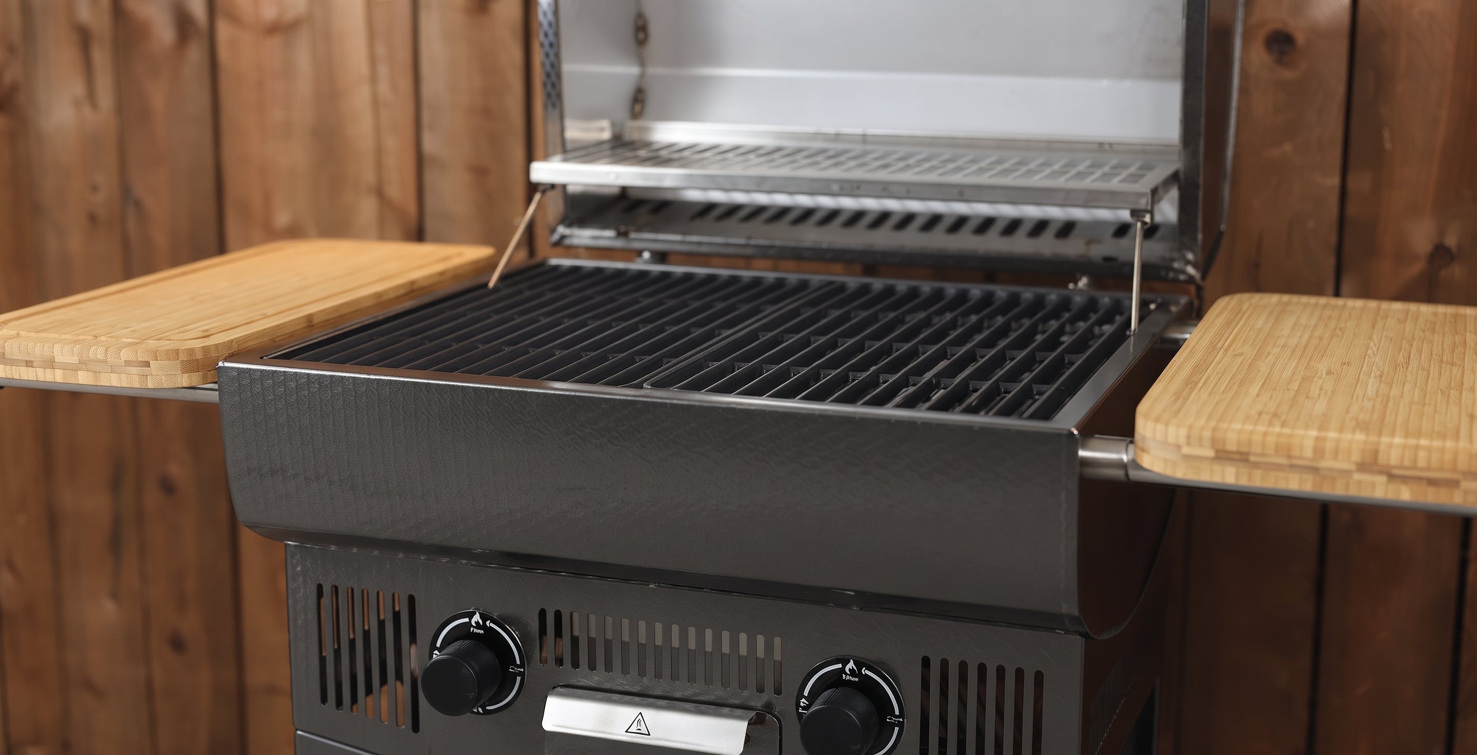 A black gas grill with a stainless steel interior, shown open with wooden side tables attached on both sides, set against a wooden plank wall.