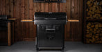 A black outdoor grill with a lid, side tables, and front control knobs, shown in a rustic indoor setting with wood-paneled walls and stacked firewood.