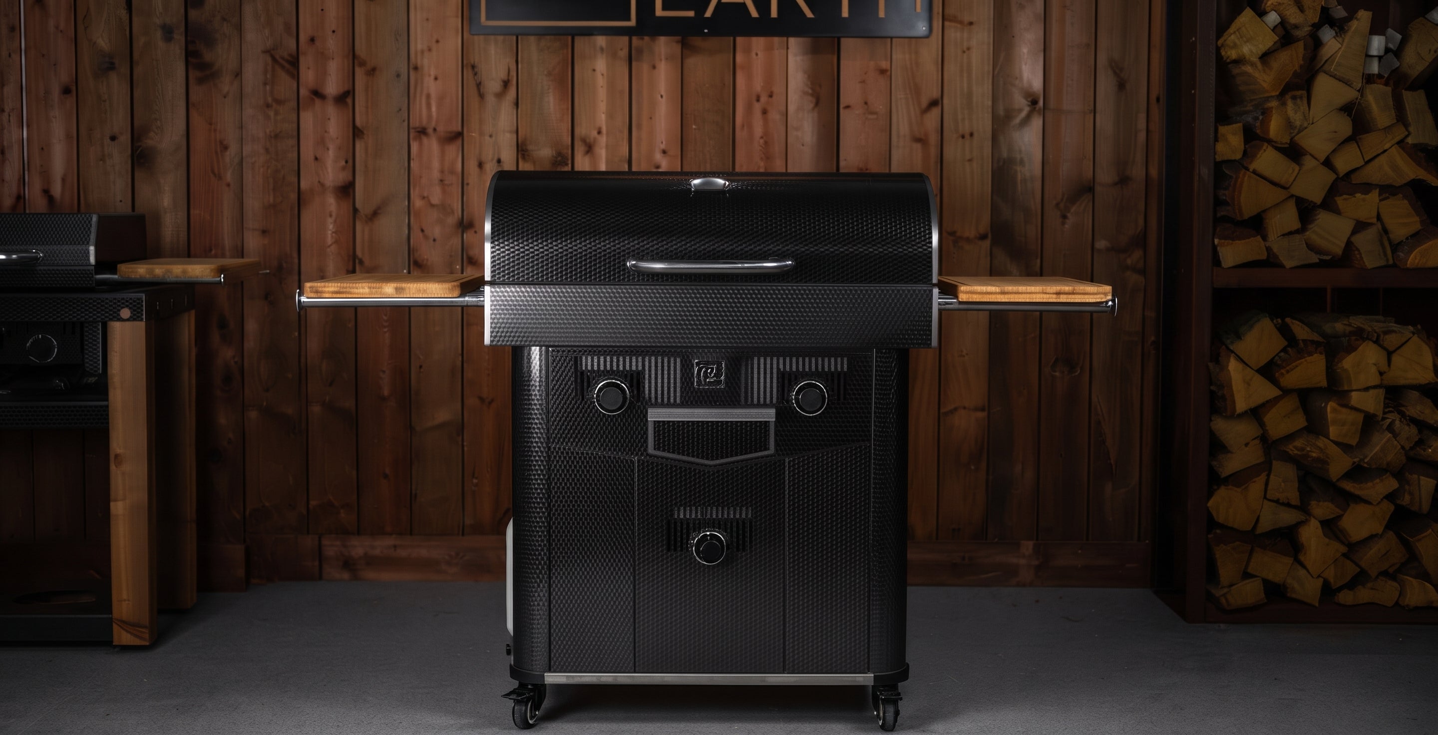 A black outdoor grill with a lid, side tables, and front control knobs, shown in a rustic indoor setting with wood-paneled walls and stacked firewood.
