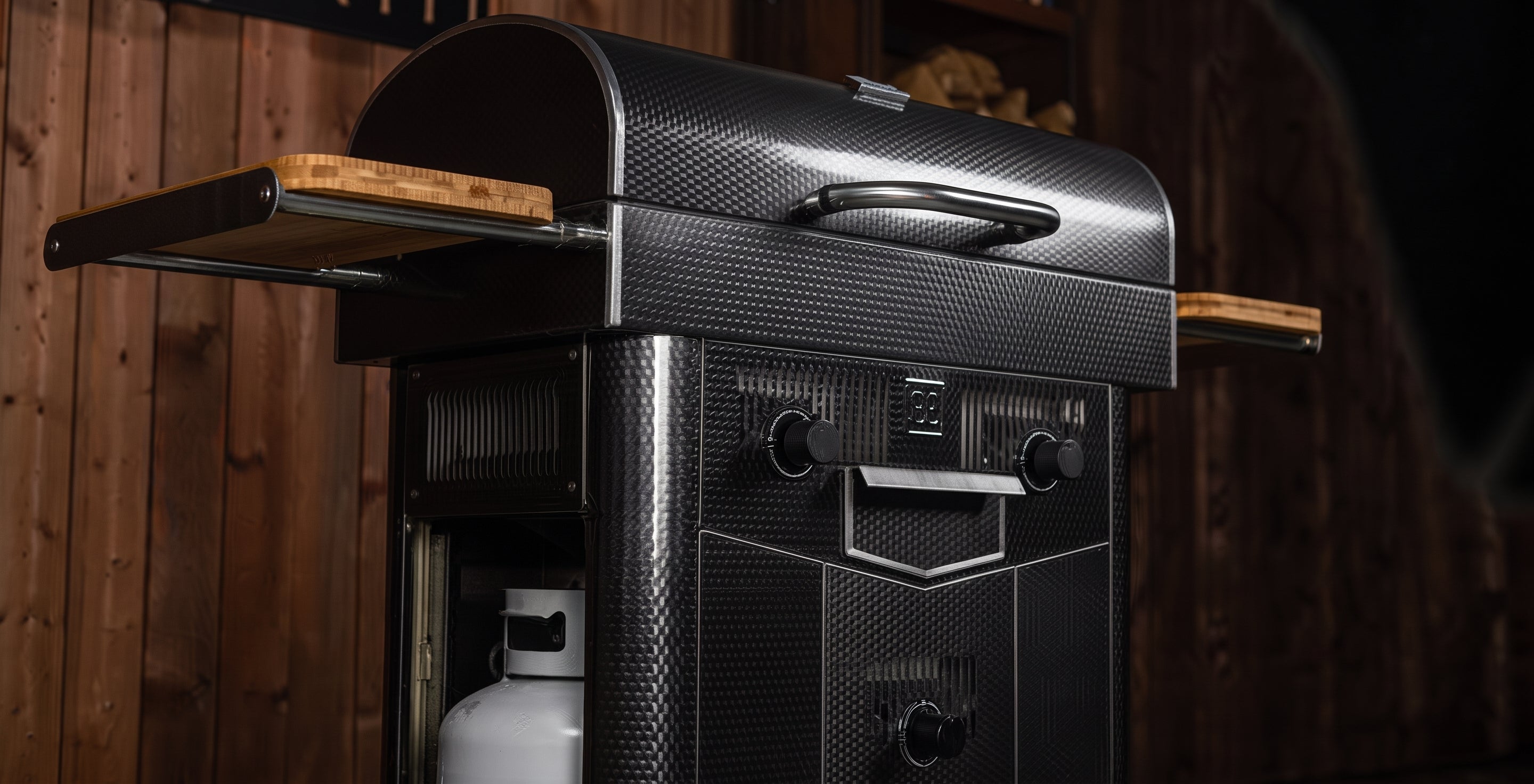 A black carbon-fiber-textured outdoor grill with wooden side shelves and a white propane tank, set against a dark wood-paneled background.