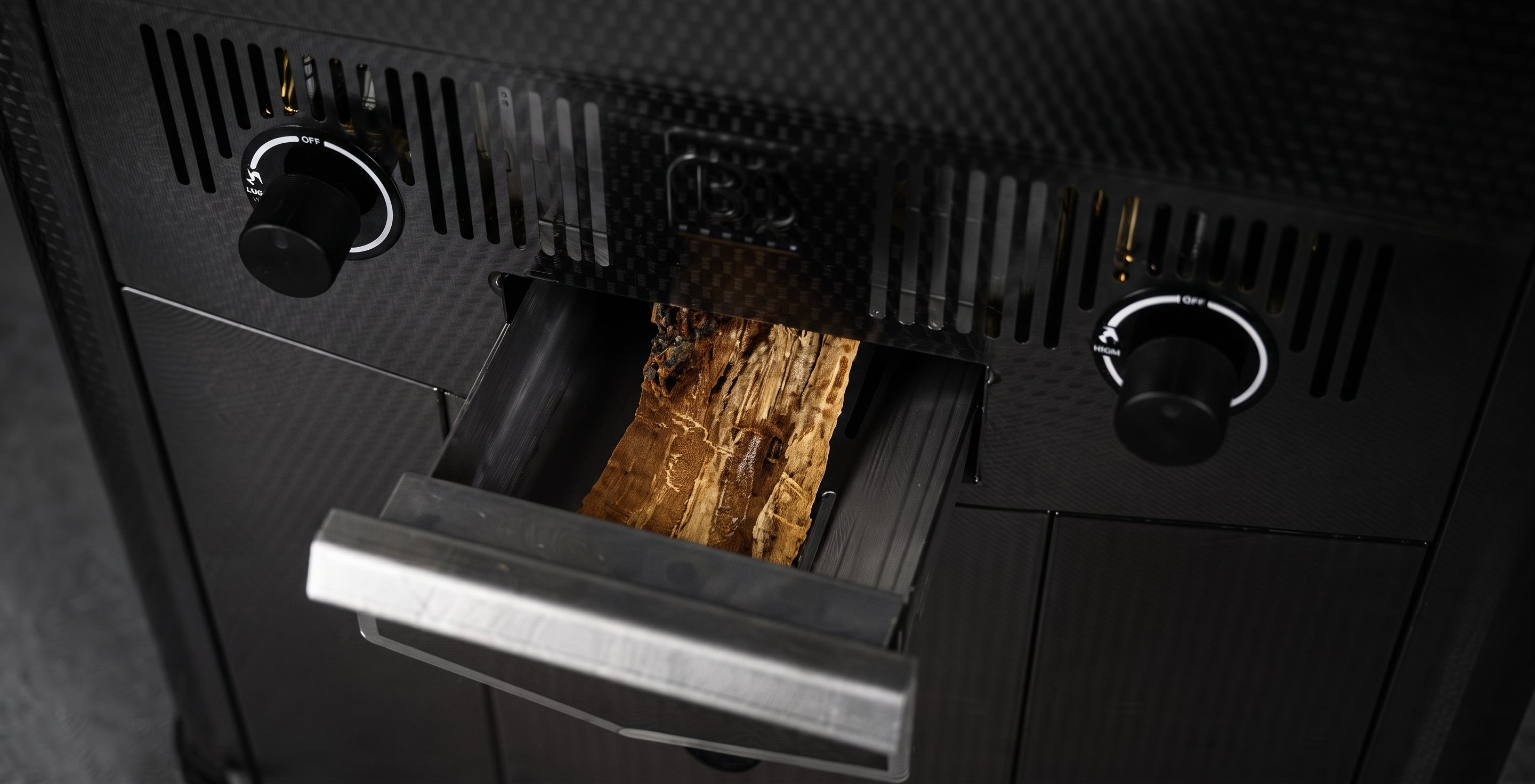 Close-up of a black wood-burning stove or fireplace insert with a pull-out door showing stacked wood logs inside, set against a dark textured background.