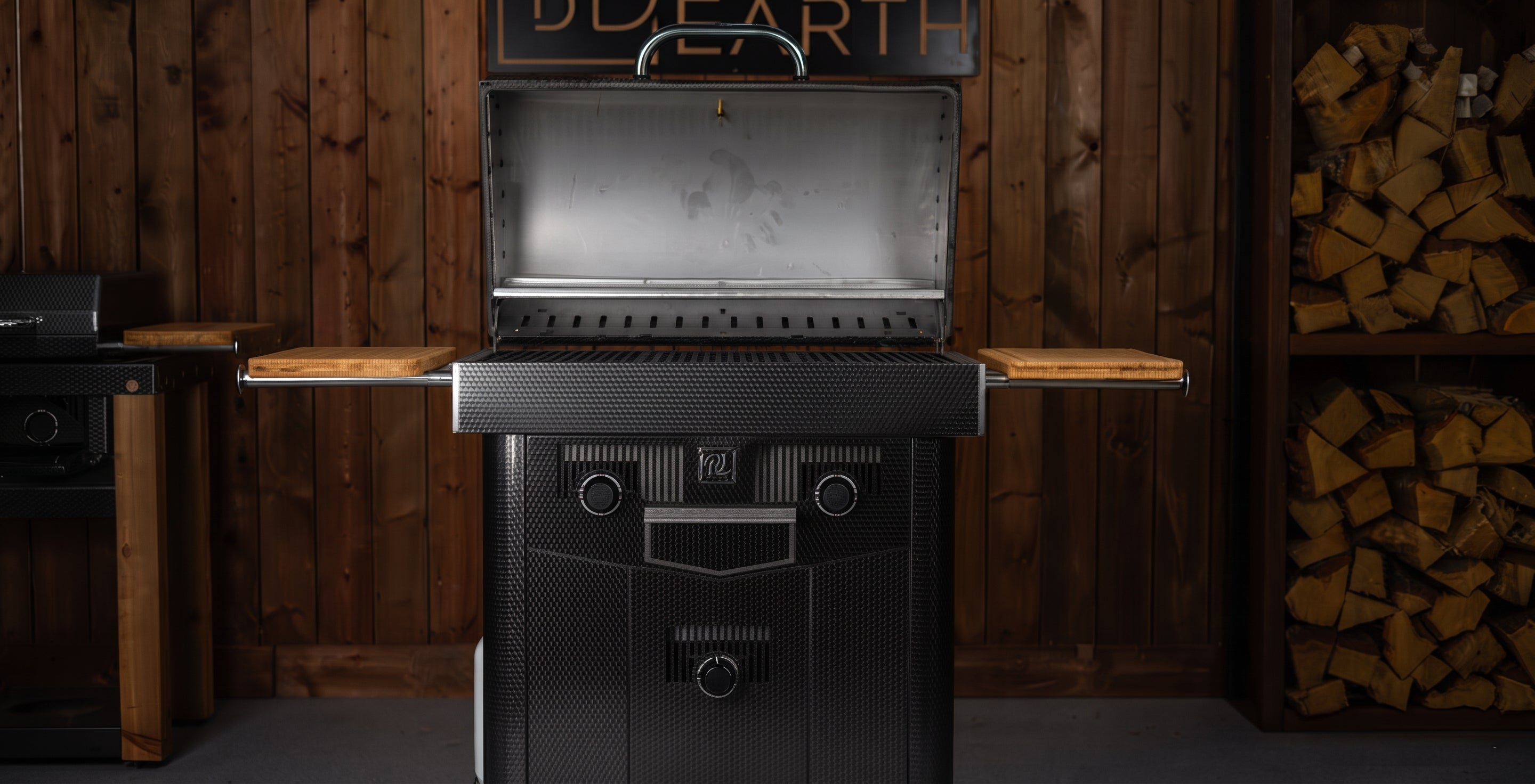 A black and stainless steel outdoor grill with wooden side tables is shown open in a rustic wood-paneled room with stacked firewood visible on the right.