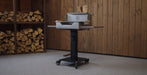 A black and silver outdoor grill on a wheeled stand, positioned in front of a wooden plank wall with stacked firewood visible on the left.