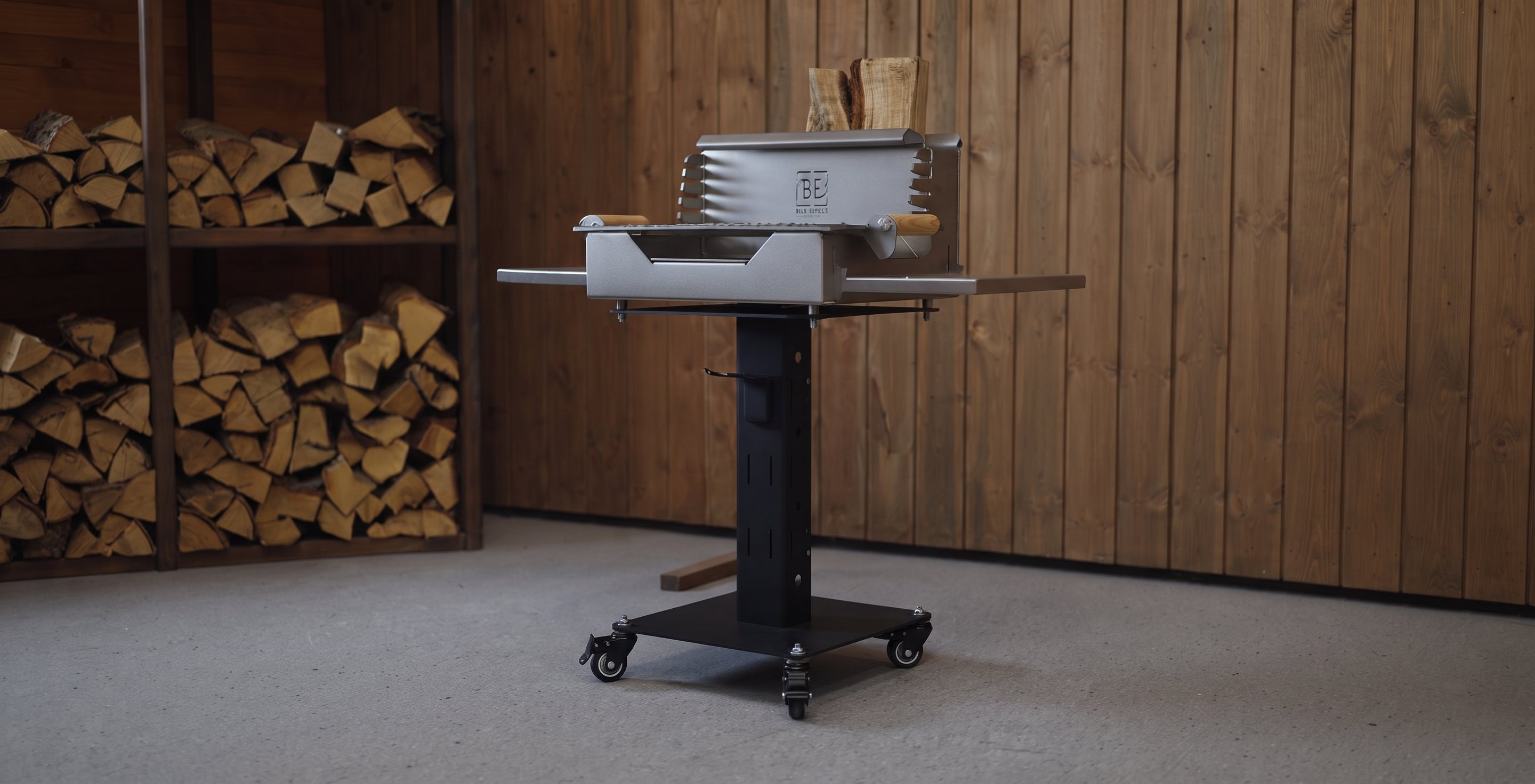 A black and silver outdoor grill on a wheeled stand, positioned in front of a wooden plank wall with stacked firewood visible on the left.
