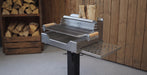 A stainless steel outdoor grill with a side shelf and wood-burning lid, shown in a rustic setting with a wood-paneled wall and stacked firewood in the background.