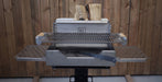 A stainless steel outdoor grill with a lid, side tables, and a wood fire basket, shown with stacked firewood in the back against a wooden fence.