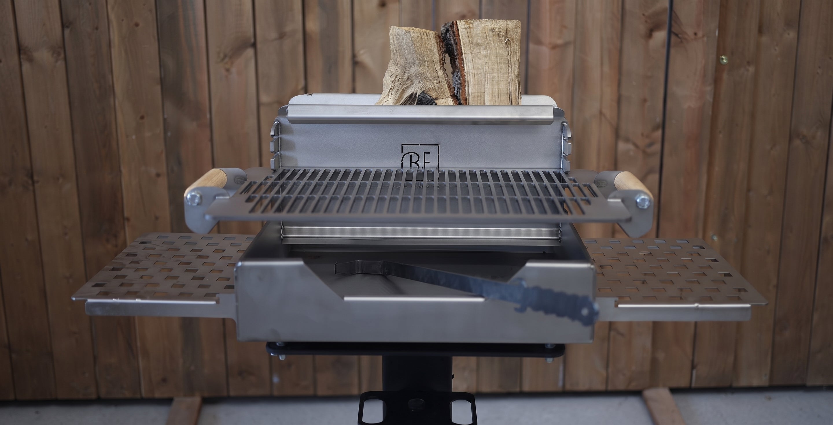 A stainless steel outdoor grill with a lid, side tables, and a wood fire basket, shown with stacked firewood in the back against a wooden fence.