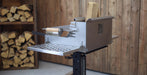 A stainless steel outdoor fire starter and wood-burning tool with a metal grate tray and a small wooden-handled tool, shown in a rustic wood-paneled room with stacked firewood in the background.