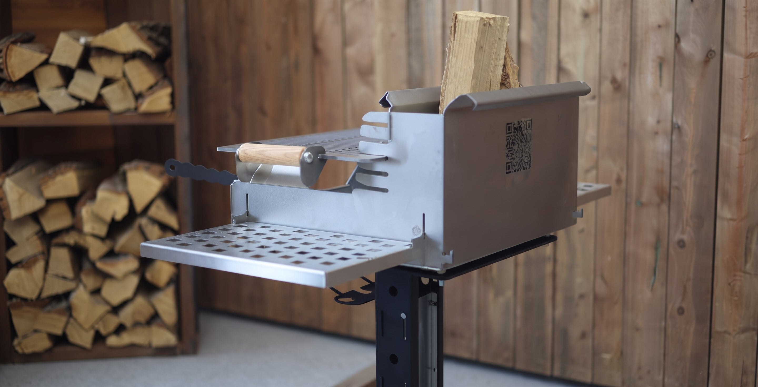 A stainless steel outdoor fire starter and wood-burning tool with a metal grate tray and a small wooden-handled tool, shown in a rustic wood-paneled room with stacked firewood in the background.