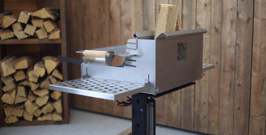A stainless steel outdoor fire starter and wood-burning tool with a metal grate tray and a small wooden-handled tool, shown in a rustic wood-paneled room with stacked firewood in the background.