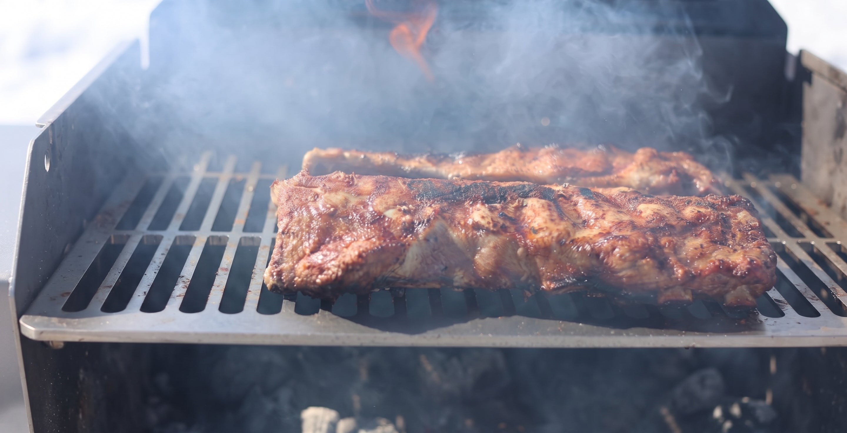 Ribs of meat cooking on a grill grate with smoke and flames visible, photographed in a real outdoor grilling environment.