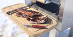 Grilled ribs resting on a wooden cutting board attached to a stainless steel outdoor grill, with a red-handled knife and tongs nearby.