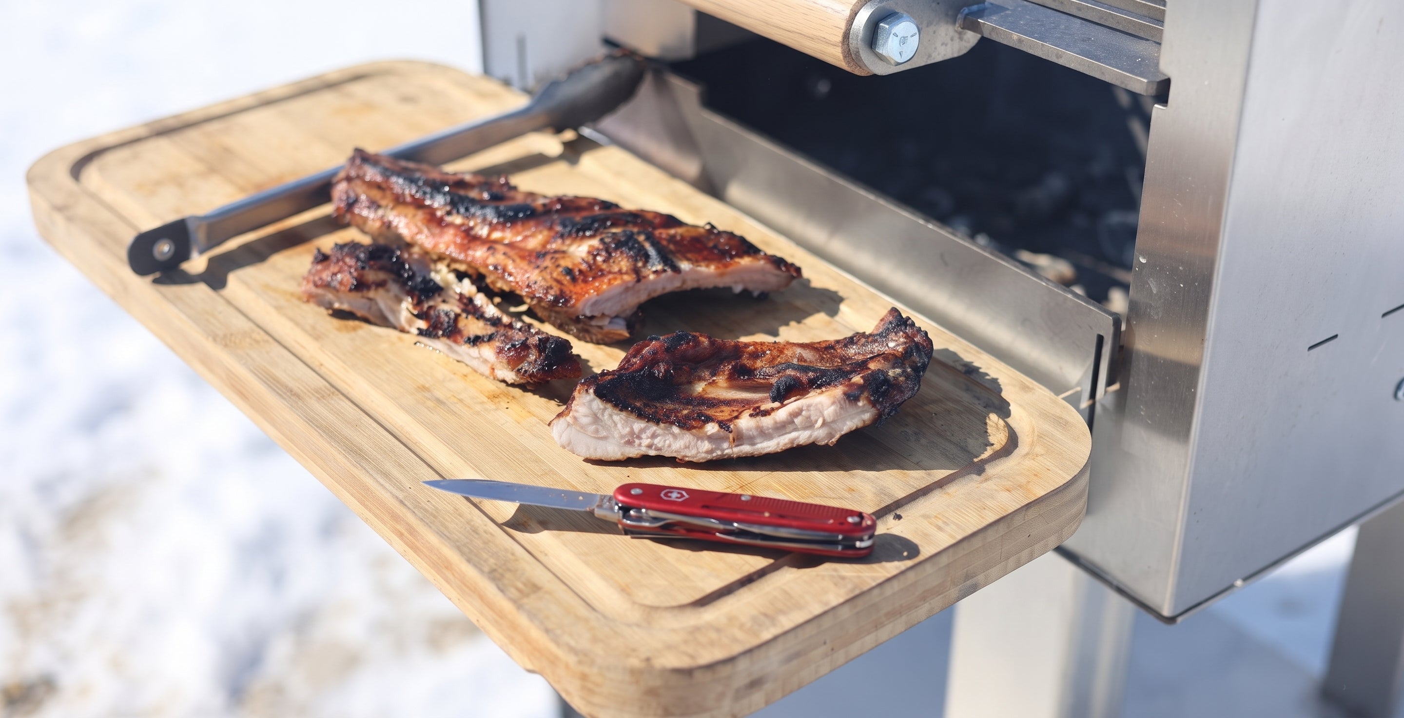 Grilled ribs resting on a wooden cutting board attached to a stainless steel outdoor grill, with a red-handled knife and tongs nearby.