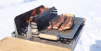 A compact metal outdoor grill with wood handles is set in snow, cooking ribs over an open flame with visible orange fire and glowing coals.