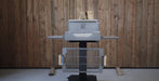 A charcoal grill with a gray metal body and black stand, positioned in front of a rustic wood-paneled wall.