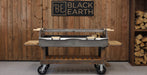 A Black Earth charcoal grill with a long spit rod and side tables, set against a rustic wood-paneled wall with a Black Earth logo sign and stacked firewood.