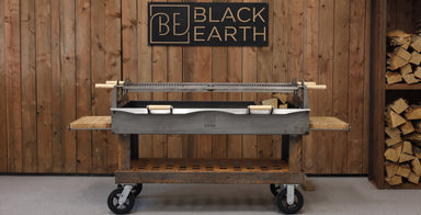 A Black Earth charcoal grill with a long spit rod and side tables, set against a rustic wood-paneled wall with a Black Earth logo sign and stacked firewood.