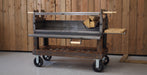 A Black Earth charcoal grill with a long spit rod and side tables, set against a rustic wood-paneled wall with a Black Earth logo sign and stacked firewood.  Open front door.