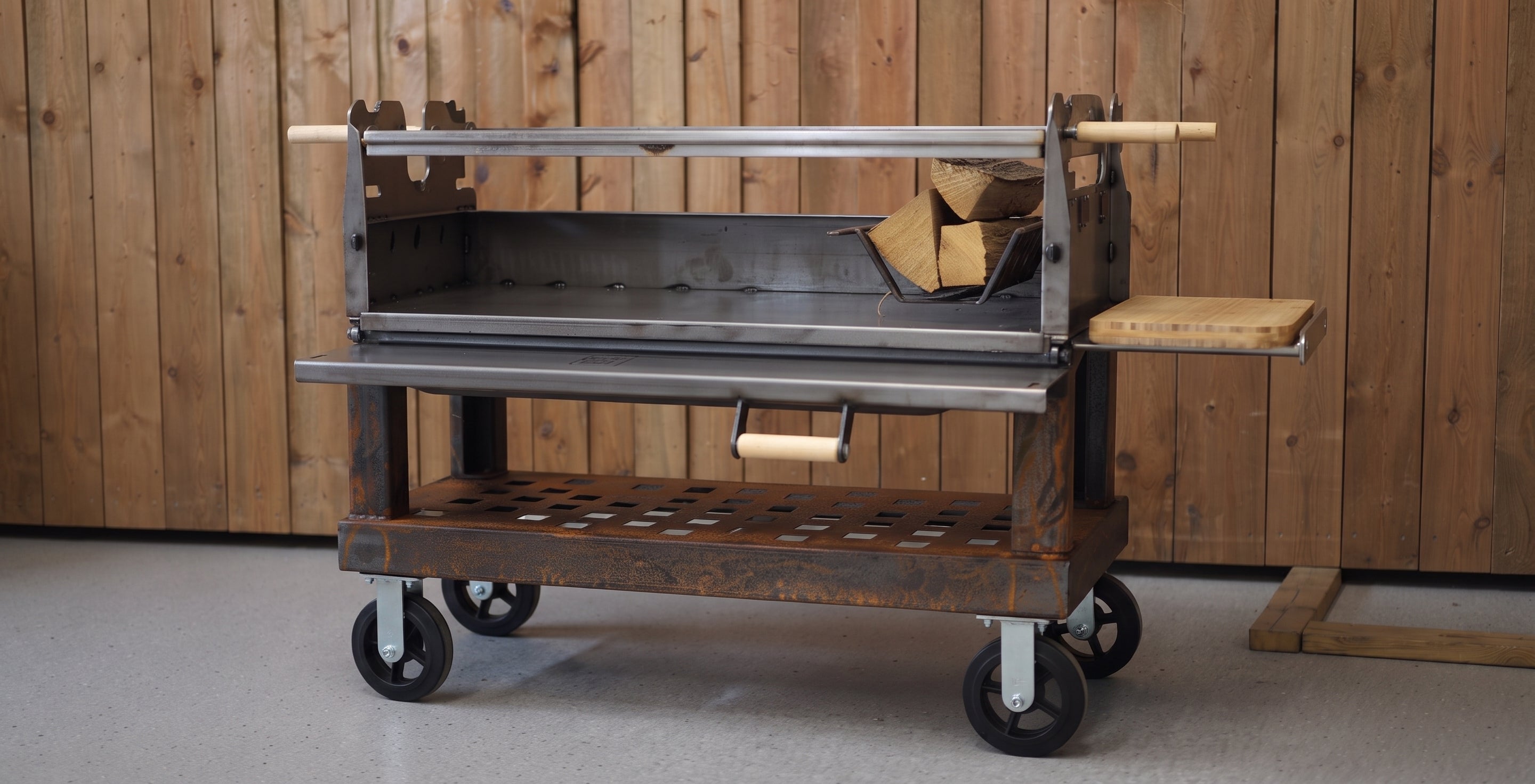 A Black Earth charcoal grill with a long spit rod and side tables, set against a rustic wood-paneled wall with a Black Earth logo sign and stacked firewood.  Open front door.