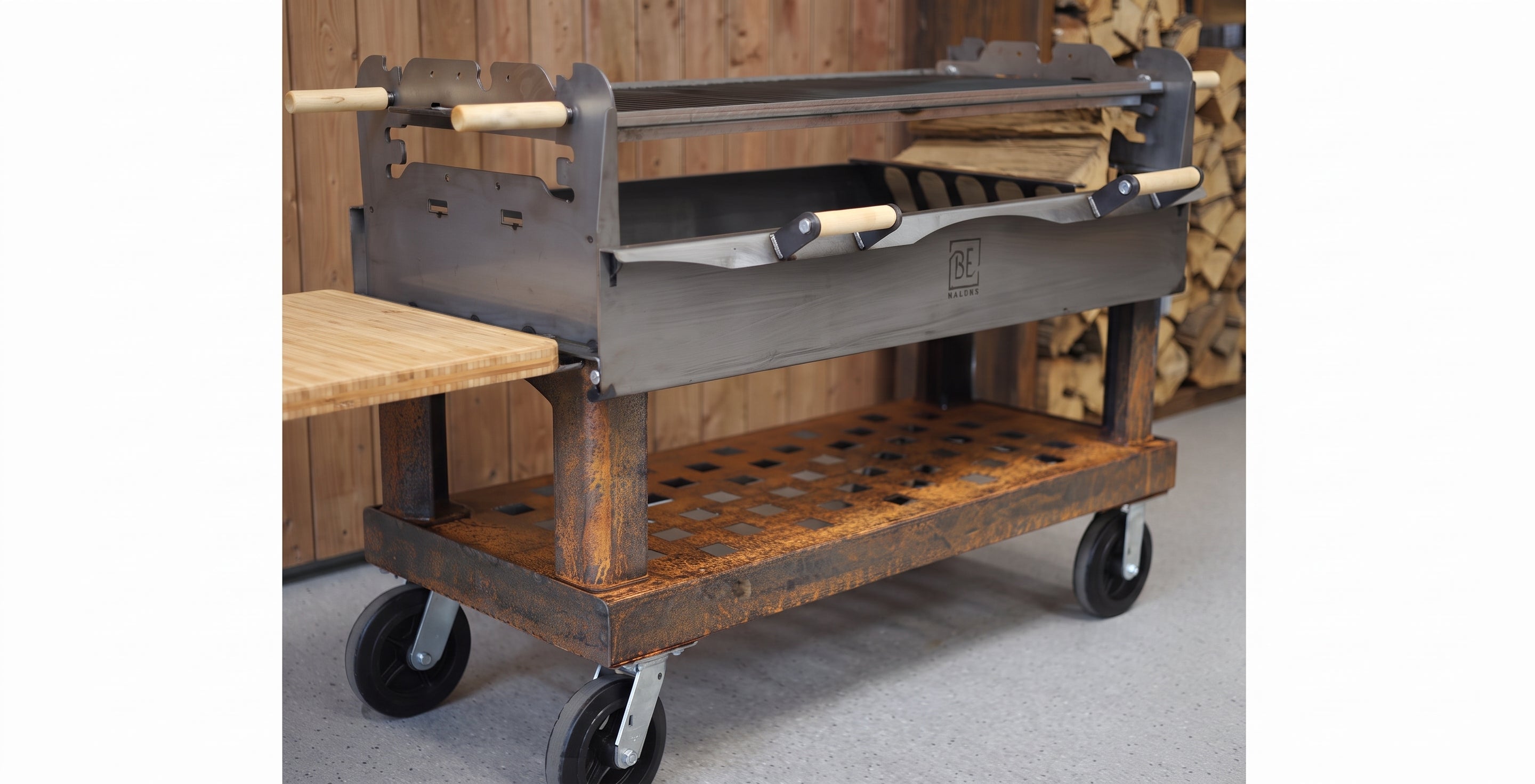 A rustic metal and wood rolling grill cart with a lower wooden shelf, black caster wheels, and a side table attachment, shown in a workshop setting with wood panels and stacked firewood in the background.