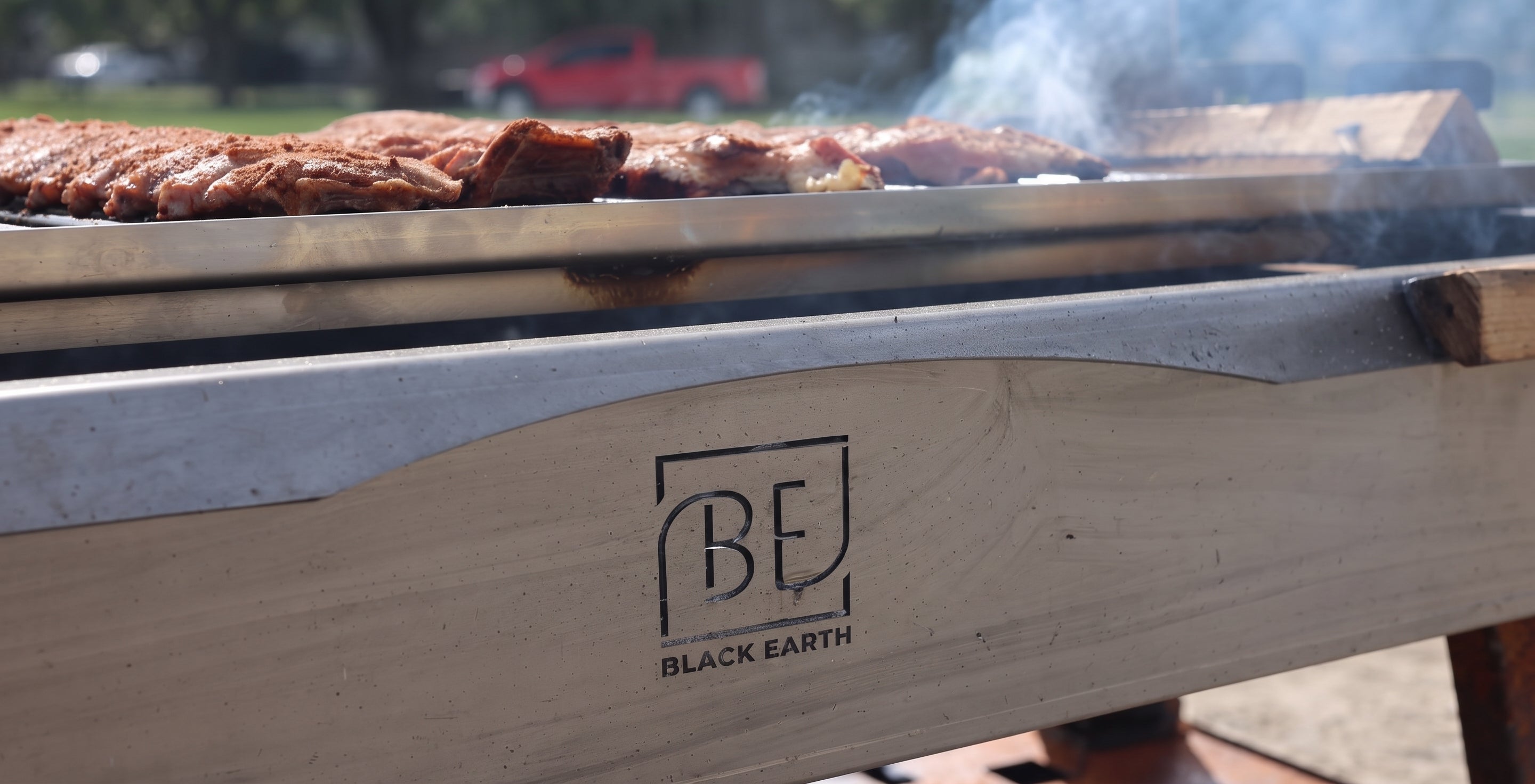 Close-up of a Black Earth outdoor grill with ribs cooking on the grate, smoke rising, and the Black Earth logo printed on the front of the wood frame.