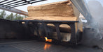 A close-up of a black metal fire starter or grill insert holding stacked wood logs with visible flames and smoke in a real outdoor setting.
