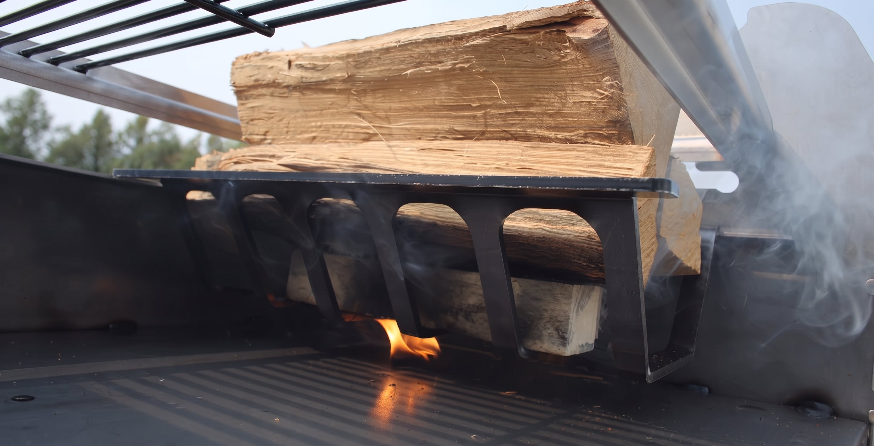 A close-up of a black metal fire starter or grill insert holding stacked wood logs with visible flames and smoke in a real outdoor setting.