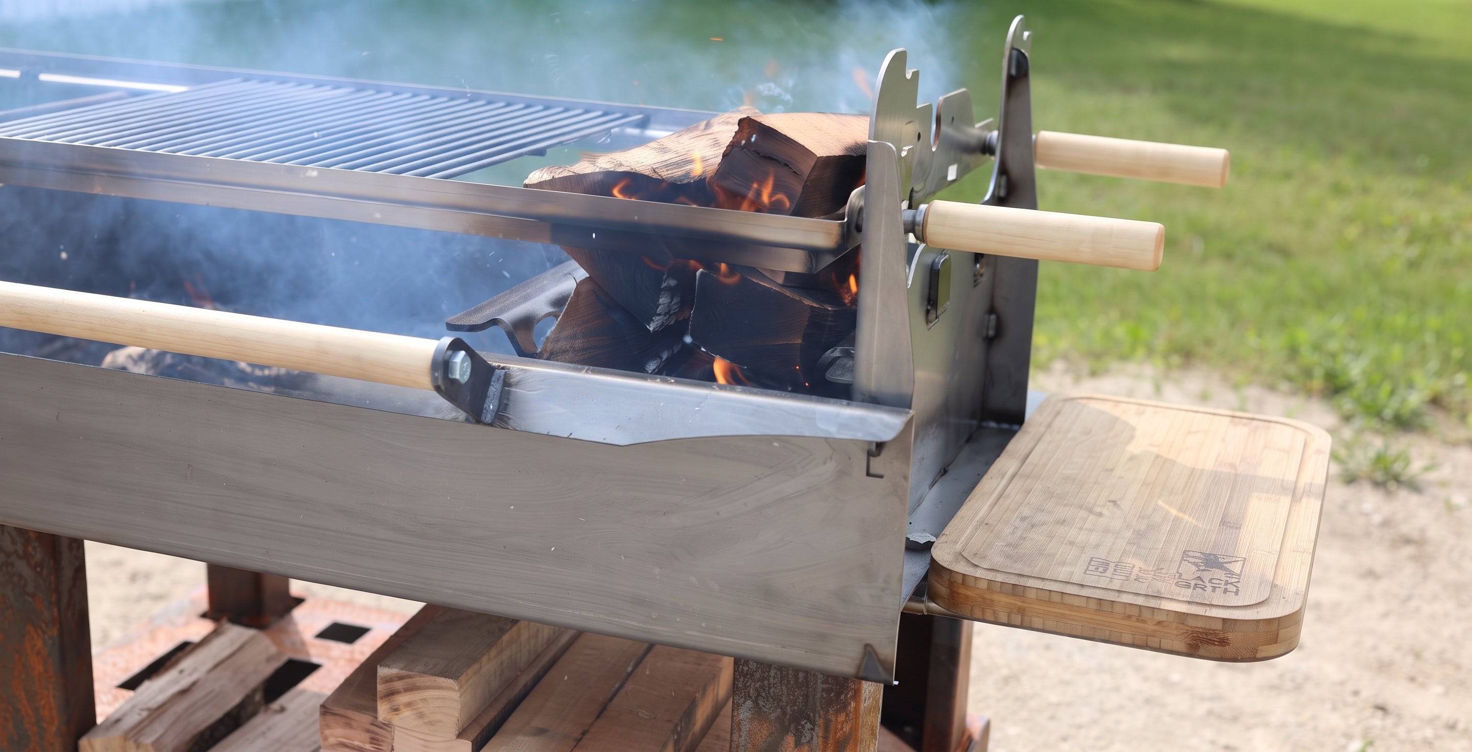 A close-up outdoor shot of a metal charcoal grill with visible flames, smoke, a wooden side table, and light-colored wooden handles on a metal frame.