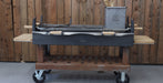 A large charcoal grill with a wooden frame and wheels, featuring a metal grate, side shelves, and a gray fuel canister, set against a wooden plank wall.