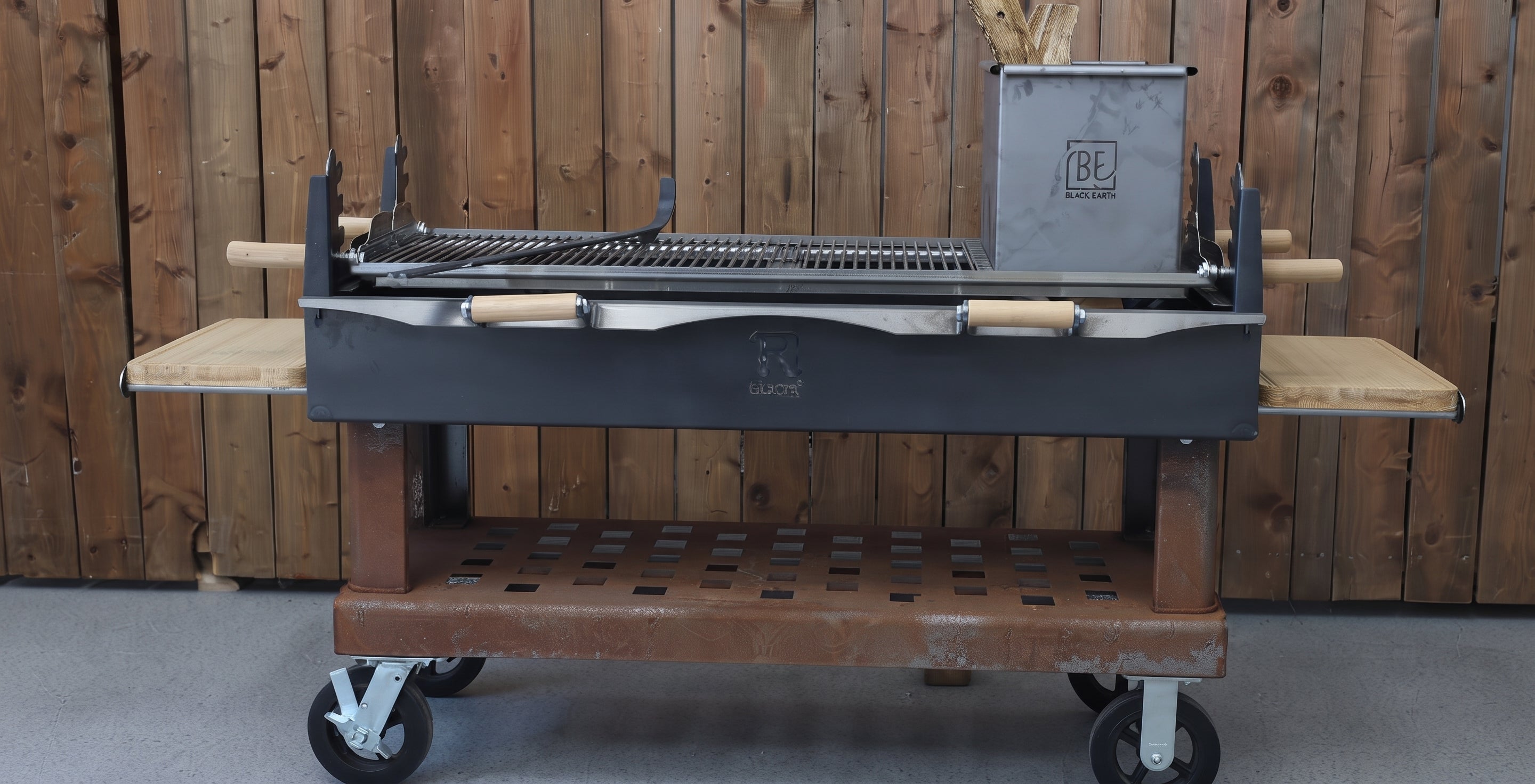 A large charcoal grill with a wooden frame and wheels, featuring a metal grate, side shelves, and a gray fuel canister, set against a wooden plank wall.