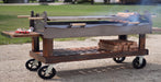 A large wheeled outdoor grill cart with a wood-grain body, side table, lower shelf with stacked wood, and black wheels, photographed outdoors on a gravel surface with grass in the background.