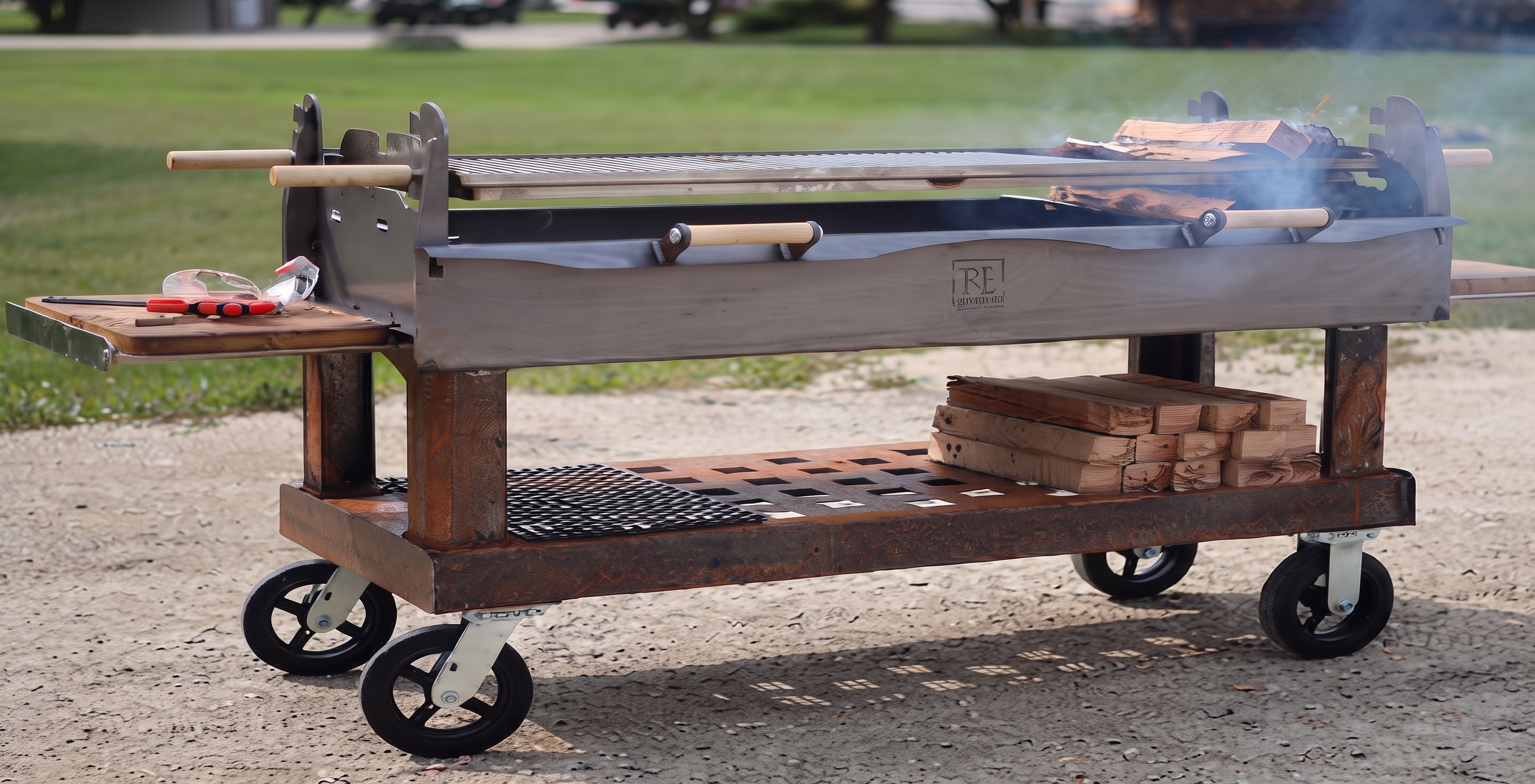 A large wheeled outdoor grill cart with a wood-grain body, side table, lower shelf with stacked wood, and black wheels, photographed outdoors on a gravel surface with grass in the background.