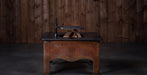 A vintage wooden table with a dark top and a carved front panel featuring a 'BE' monogram, holding a small black and brass object, set against a dark wood plank wall.