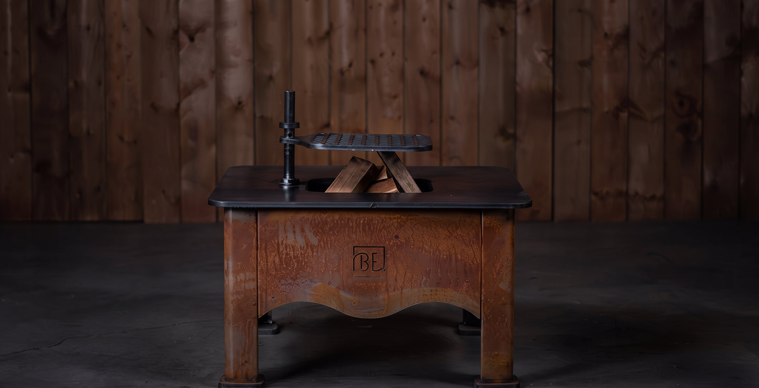 A vintage wooden table with a dark top and a carved front panel featuring a 'BE' monogram, holding a small black and brass object, set against a dark wood plank wall.