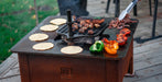 A portable outdoor grill with a dark metal cooking surface sits on a wooden deck. Tortillas, mushrooms, meat, sliced red peppers, and half green and red bell peppers are cooking on the grill, with tongs holding food above the surface.