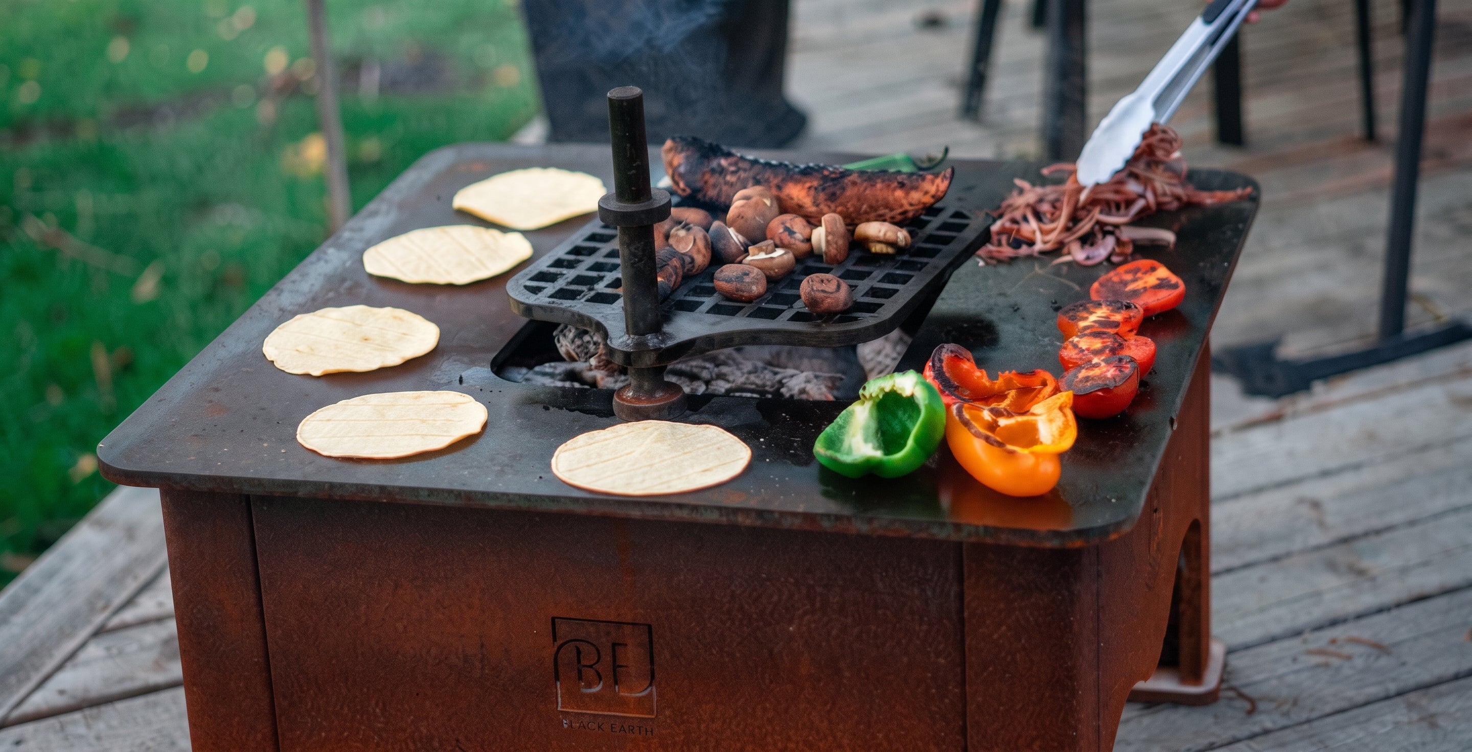 A portable outdoor grill with a dark metal cooking surface sits on a wooden deck. Tortillas, mushrooms, meat, sliced red peppers, and half green and red bell peppers are cooking on the grill, with tongs holding food above the surface.