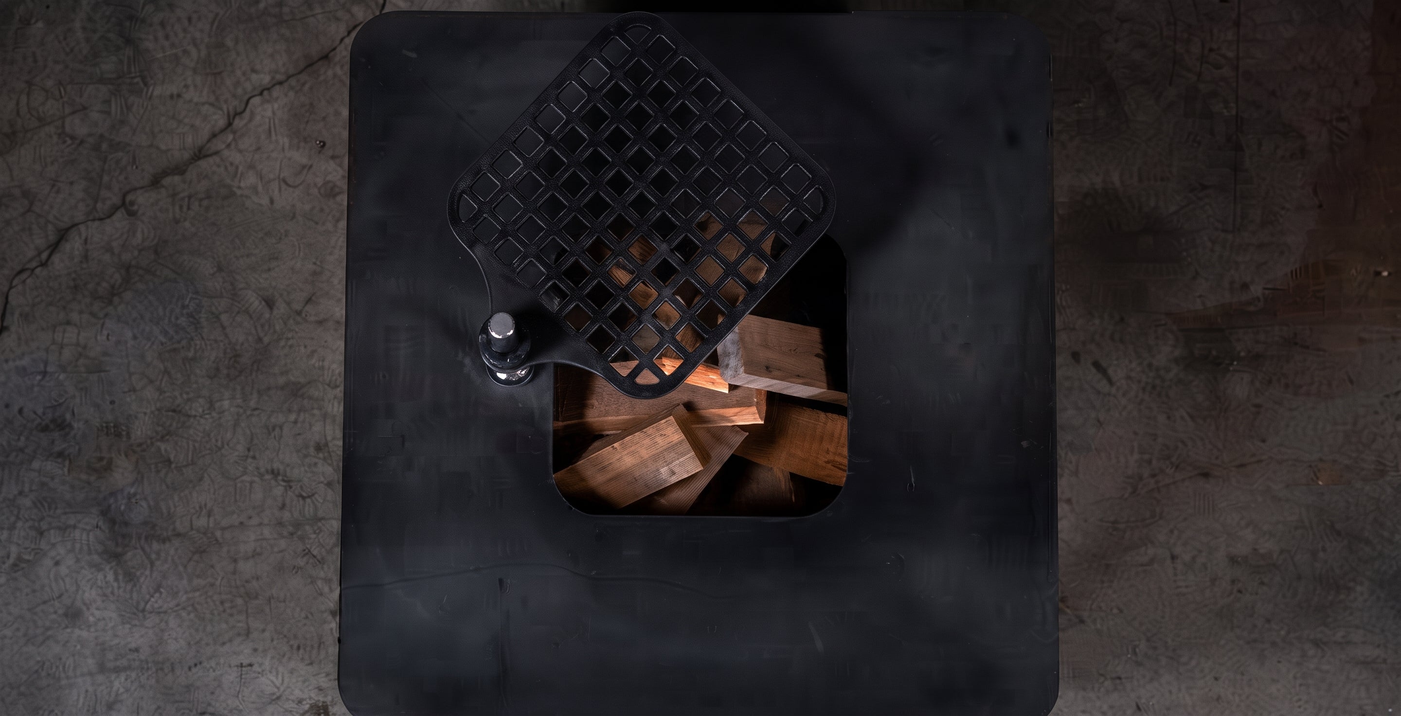 Top-down view of a black metal fireplace insert with a diamond-patterned lid and visible wood logs inside, set on a dark textured floor.