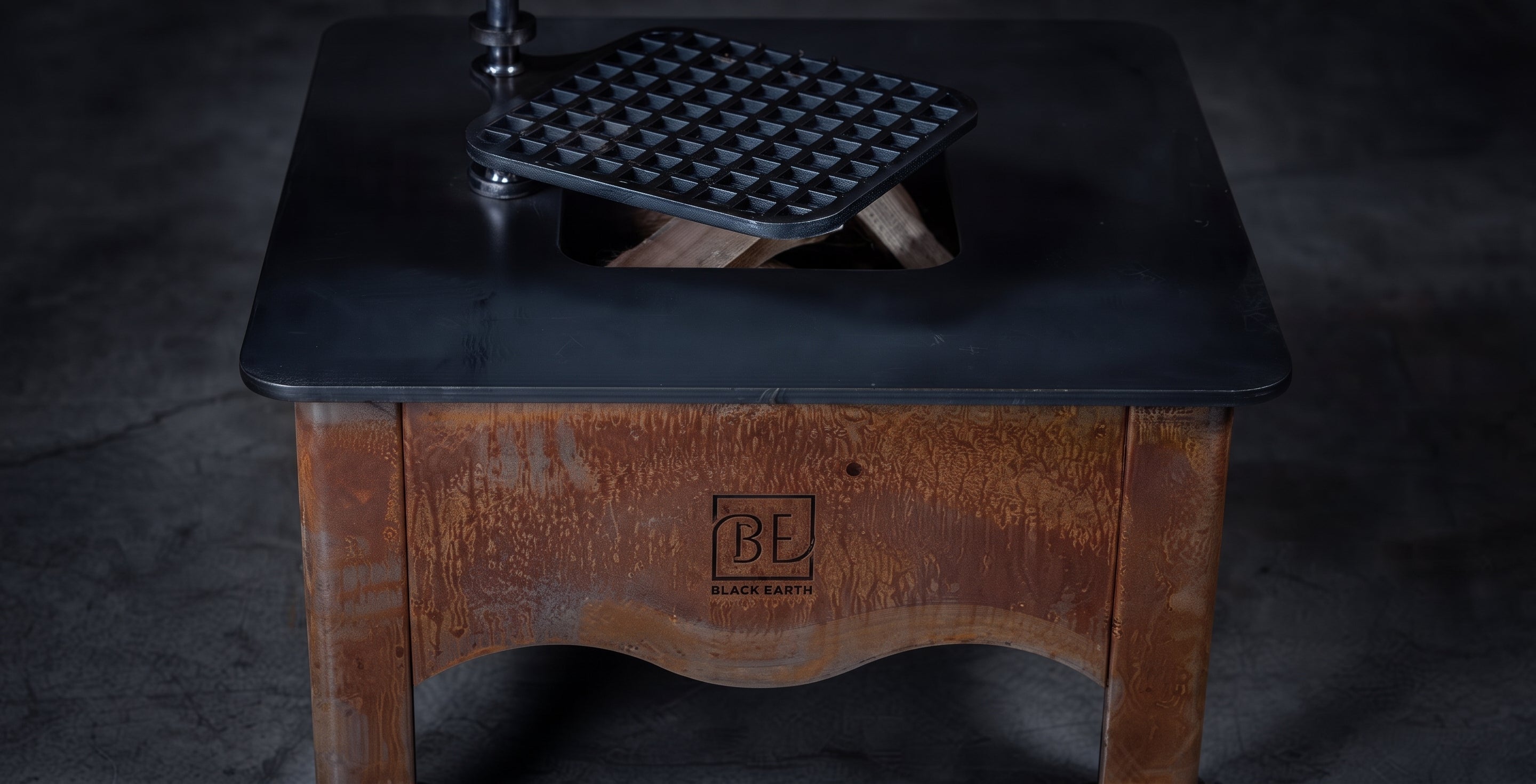 A rustic wooden table with a black metal top featuring a built-in grill or waffle maker insert, shown on a dark concrete floor. The front of the table has a carved logo that reads 'BE BOSTON EATERY'.