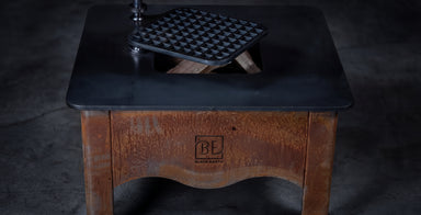A rustic wooden table with a black metal top featuring a built-in grill or waffle maker insert, shown on a dark concrete floor. The front of the table has a carved logo that reads 'BE BOSTON EATERY'.