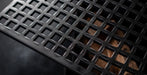 Close-up of a black metal table or floor grate with a square grid pattern, showing a brown surface underneath through the openings.
