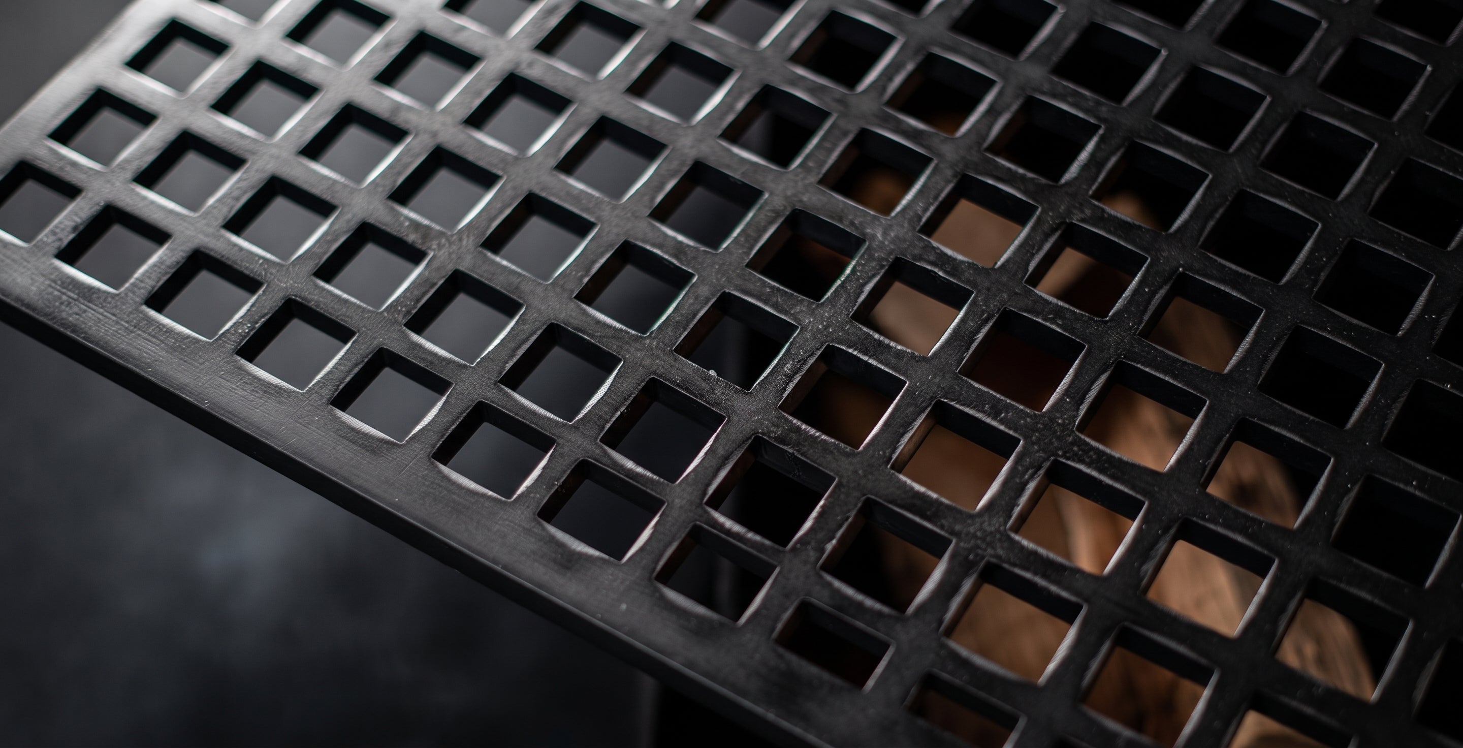 Close-up of a black metal table or floor grate with a square grid pattern, showing a brown surface underneath through the openings.