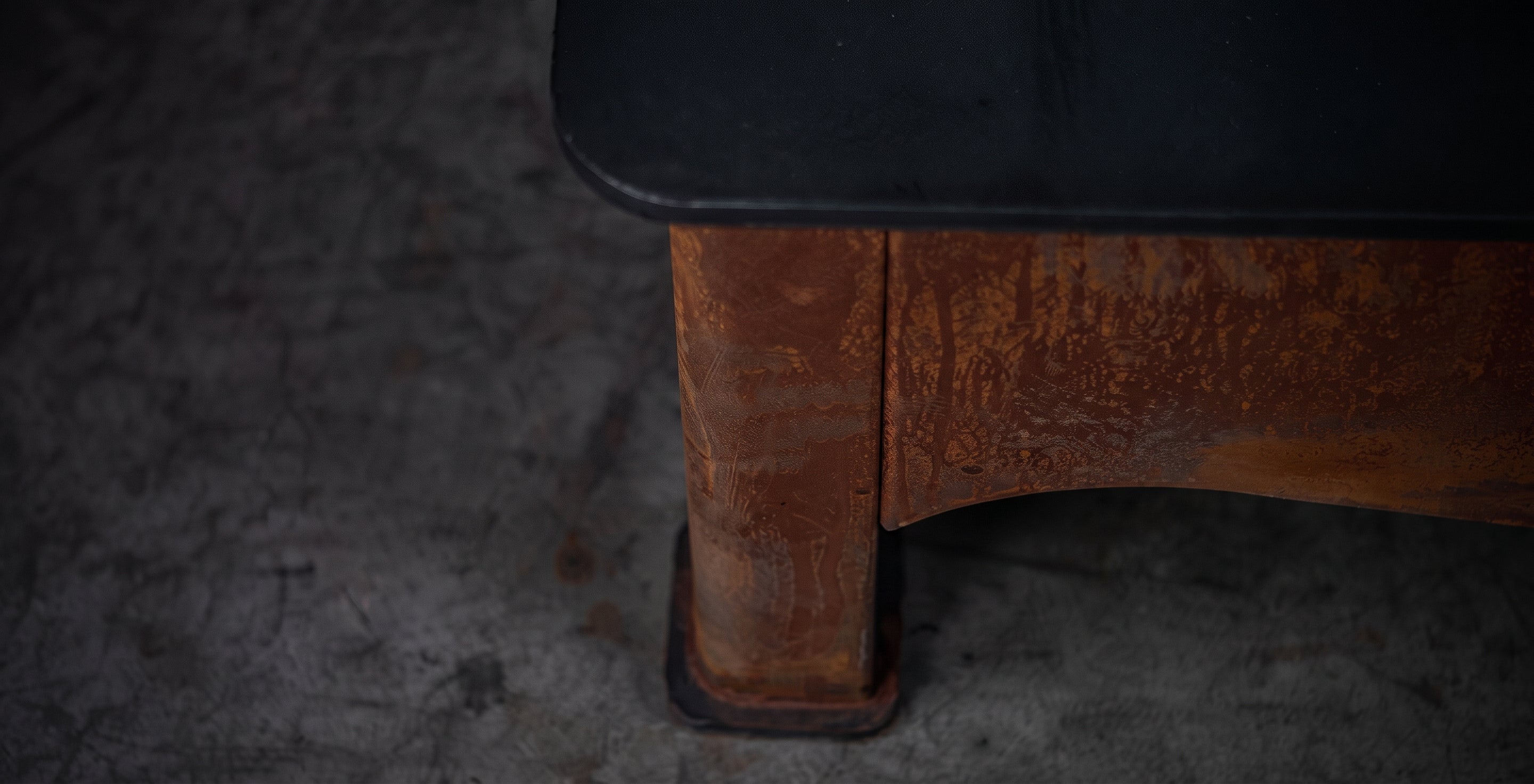 Close-up of a rustic wooden table leg with a dark top surface, set against a dark textured concrete floor.