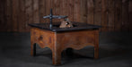A rustic wooden fire pit table with a dark metal top and a black metal wind guard, set against a dark wood plank wall.