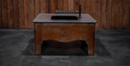 A rustic brown wooden outdoor table with a black sink basin and faucet, set against a dark wood plank wall and concrete floor.