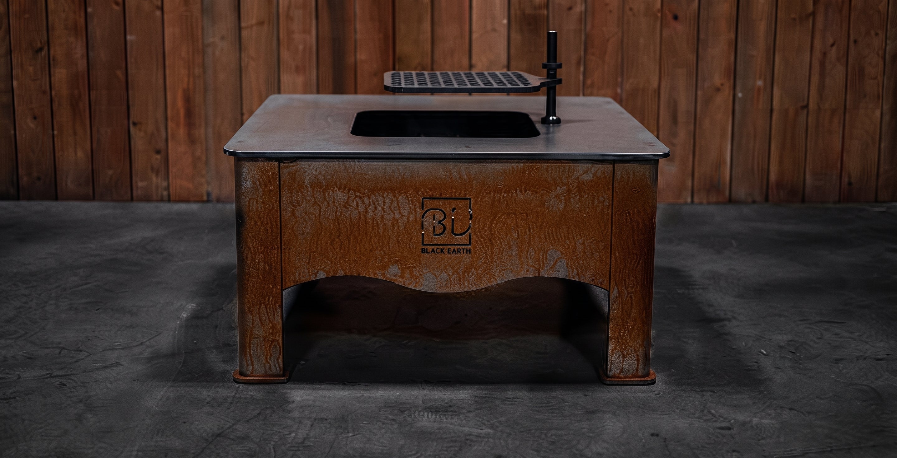 A rustic brown wooden outdoor table with a black sink basin and faucet, set against a dark wood plank wall and concrete floor.