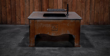 A rustic brown wooden outdoor table with a black sink basin and faucet, set against a dark wood plank wall and concrete floor.